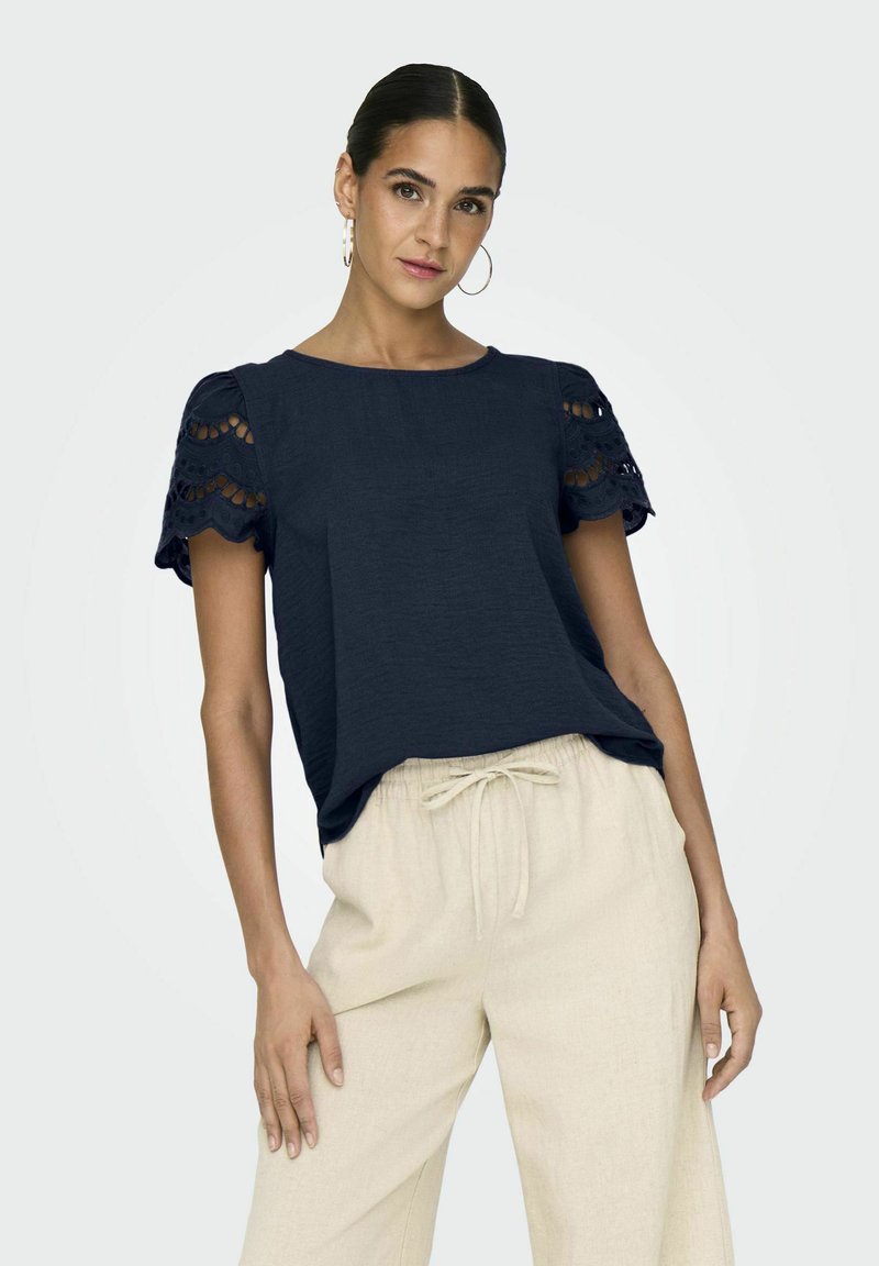 Haut bleu marine à manches courtes avec des détails en dentelle aux épaules, coupe décontractée, associé à un pantalon beige clair à cordon de serrage. Texture lisse.