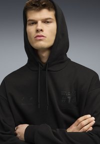 Jonge man met kort haar, gekleed in een zwarte hoodie met BMW Motorsport-logo's, met zijn armen over elkaar, tegen een grijze verloop achtergrond.