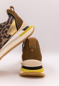 Bruna och leopardmönstrade sneakers med gulddetaljer och lager av gula, svarta och beiga sulor på en enfärgad ljus bakgrund.