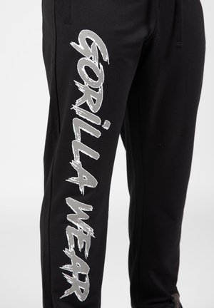 Pantalon de survêtement noir en tissu doux, arborant un grand texte argenté "GORILLA WEAR" sur le côté. Taille élastique avec cordon de serrage pour un ajustement personnalisé.
