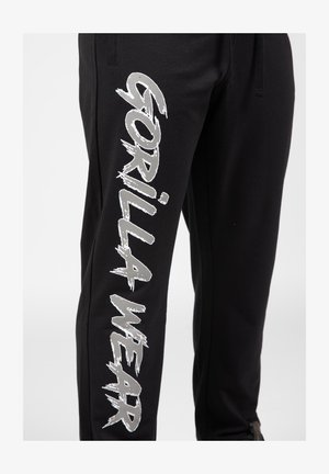 Zwarte sweatpants van zacht materiaal, met grote zilveren 'GORILLA WEAR' tekst aan de zijkant. Elastische tailleband met trekkoord voor aanpassing.