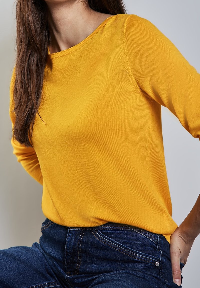 Femme portant un haut à manches longues jaune vif associé à un jean bleu foncé, posant avec une main sur la hanche devant un fond neutre.