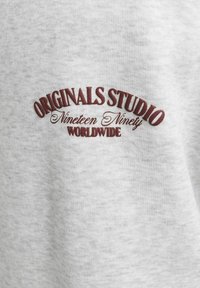 Tela de sudadera gris con una superficie texturizada. El texto bordado en rojo dice "ORIGINALS STUDIO" sobre "Nineteen Ninety" y "WORLDWIDE."