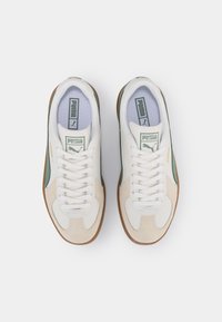 Paire de baskets blanches et beiges Puma avec des accents verts et des semelles en gomme, vues de dessus sur fond blanc uni.