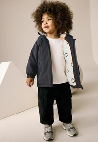 Enfant portant une veste zippée grise foncé avec une doublure à motifs, une chemise blanche, un pantalon cargo noir et des baskets grises, posé contre un fond neutre.
