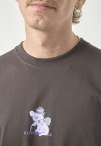 Dark gray short-sleeve t-shirt featuring a purple and green cartoon dinosaur graphic on the chest, with text below reading "CLEPTOMANIX."