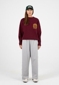 Burgundowy, oversize sweter z złotym emblematem, w połączeniu z jasnoszarymi spodniami o szerokich nogawkach z bocznymi paskami oraz białymi trampkami.
