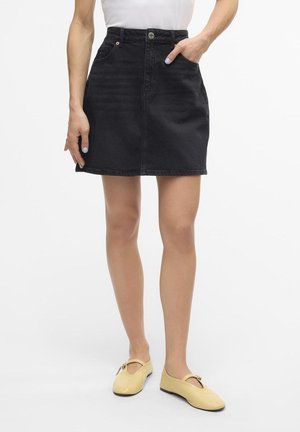 Vrouw die een zwarte denim minirok met voorzakken draagt en beige Mary Jane schoenen aan heeft, staand met één hand in de zak.