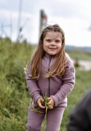 Kind trägt ein helllila Fleece-Outfit, hält in beiden Händen Gras. Lange braune Haare, draußen mit grüner Umgebung im Hintergrund.