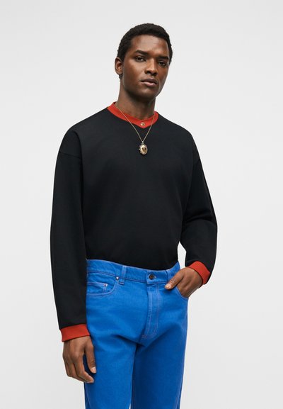 Νέος άνδρας με σκούρο δέρμα που φοράει μαύρο μακρυμάνικο πουκάμισο με κόκκινες μανσέτες, φωτεινό μπλε τζιν και κολιέ με μενταγιόν, μπροστά σε απλό φόντο.