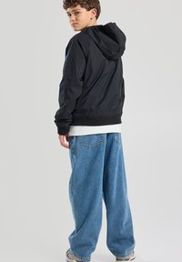 Mustat hupparitakki, jossa on sileä pinta, yhdistettynä volyymiltaan väljiin sinisiin farkkuihin. Huomionarvoisia yksityiskohtia ovat valkoinen aluspaidat ja lenkkarit.