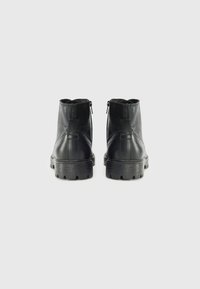 Botines de cuero negro con una suela gruesa, mostrados desde la parte trasera, con cremalleras en los costados y un diseño minimalista sobre un fondo blanco.