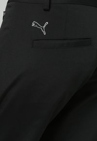 Svarta byxor i ett slätt tyg, med en sydd horisontell ficka och en vit Puma-logga broderad nära fickan.