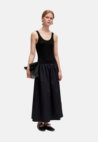 Zwarte mouwloze top met geribbeld textuur, gecombineerd met een vloeiende zwarte satijnen rok. Houdt een zwarte clutch vast en draagt zwarte veterschoenen.