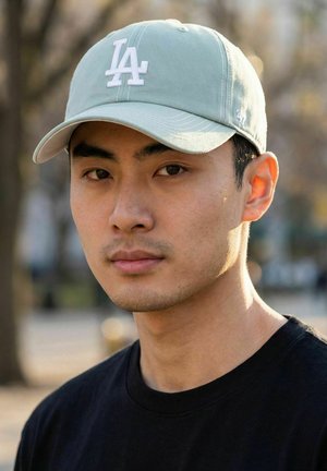 Young man wearing a light gray LA baseball cap and black shirt, looking directly at camera with blurred outdoor background.