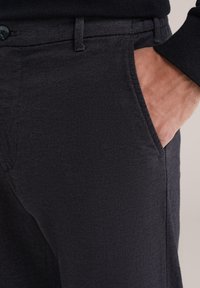 Ruka muže spočívající v přední kapse tmavých, texturovaných kalhot, na sobě má viditelný rukáv černého svetru.