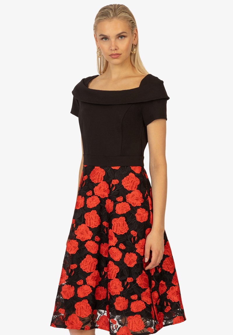 Schwarzes Kleid mit eng anliegendem Oberteil und schulterfreiem Ausschnitt, ausgestattet mit einem schwingenden Rock, der mit roten Rosenapplikationen auf schwarzer Spitze verziert ist.