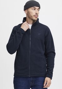 Chaqueta de forro polar azul marino con cuello alto, cremallera completa y bolsillos laterales. Presenta una textura suave y un logo sutil en el pecho. Combinada con vaqueros azules.