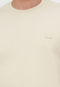 Μπλουζάκι σε κρεμ απόχρωση με στρογγυλή λαιμόκοψη και ένα μικρό τετράγωνο φιλέτο στο στήθος που απεικονίζει το όνομα "Pier One".