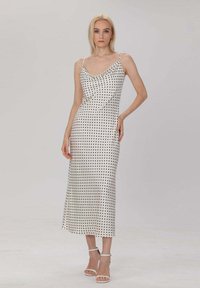 Robe mi-longue à pois en blanc et noir, avec un col drapé, des fines bretelles et une texture satinée lisse. Complétée par des talons blancs.