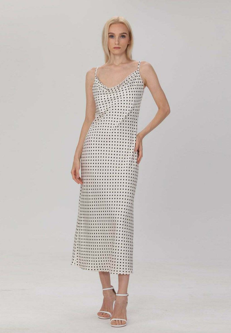 Robe mi-longue à pois en blanc et noir, avec un col drapé, des fines bretelles et une texture satinée lisse. Complétée par des talons blancs.