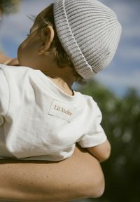 Vit bomulls t-shirt med en beige etikett som står "Lil'Atelier" på baksidan; barn som bär en ljusgrå stickad mössa.