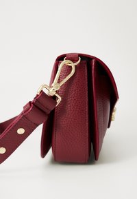 Dark red textured leather crossbody bag with a wide strap attached by gold clasp and rivets, shown from side on a white background.