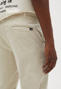 Primo piano di una persona che indossa pantaloni beige e una maglietta bianca con testo nero, con la mano appoggiata di lato.