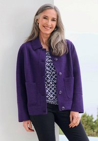Giacca di lana viola con due tasche frontali, chiusura a bottoni e colletto, indossata sopra una blusa grigia a fantasia e pantaloni neri.