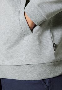 Sudadera de color gris claro con una tela texturizada, bolsillo canguro y puños acanalados. Presenta una pequeña etiqueta de la marca en el lateral.