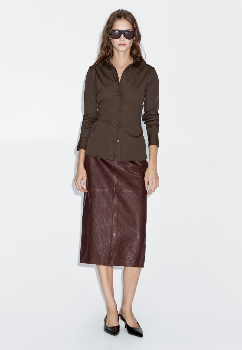 Bruine blouse van gladde stof, gecombineerd met een leren kokerrok in bruin. Model draagt zwarte zonnebril en puntige zwarte hakken.