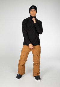 Maglietta nera a maniche lunghe, pantaloni cargo marroni con tasche laterali, berretto nero e scarpe nere con suole bianche. Sfondo semplice.