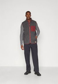 Siv jopič iz flisa s bordo prsnim žepom, z zadrgami in prilegajočo se zasnovo, kombiniran z temnimi hlačami in črnim čevljem.