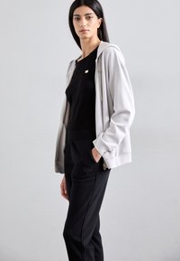 Sweat à capuche zippé gris clair avec cordons, t-shirt noir ajusté en dessous, et joggers noirs avec logo sur la poche, présentés contre un arrière-plan neutre.