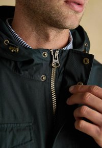 Jachetă verde închis cu fermoar frontal, accente aurii și glugă cu șnur. Are un guler în dungi și detalii cu nasturi în zona pieptului.