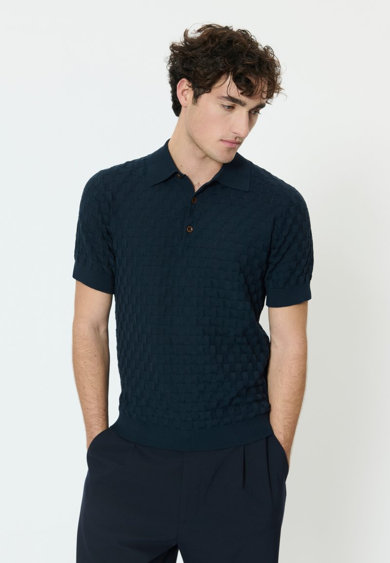 Mladi moški s kodrastimi lasmi, oblečen v temno modro teksturirano polo majico s kratkimi rokavi in temne hlače, gleda navzdol z rokami v žepih.