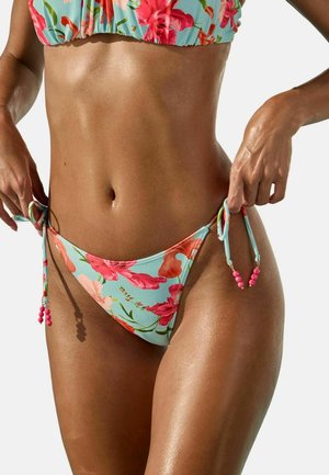 Femeie purtând un bikini floral albastru, ajustând șnururile din părțile laterale ale slipului de bikini, arătând abdomenul și brațele tonifiate.