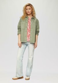 Olivgrüne Jeansjacke über einem pinkfarbenen gemusterten Top, kombiniert mit hellblauen Schlagjeans und beigen Sneakers. Texturiertes, lässiges Design.