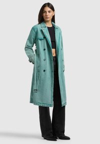 Giovane donna che indossa un lungo trench doppiopetto verde acqua, top corto nero, jeans neri e scarpe nere con punta, in piedi con le mani nelle tasche.