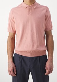 Uomo che indossa una polo a maniche corte di maglia rosa chiaro con orlo a costine e pantaloni neri, su uno sfondo bianco semplice.