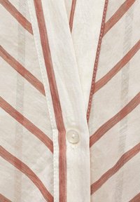 Camicia bianca con righe diagonali rosse. Tessuto leggero, cuciture visibili e un bottone rotondo al colletto.