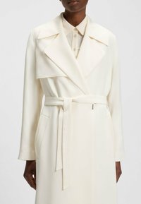Cremfarbener, taillierter Trenchcoat mit weitem Kragen, strukturierten Schultern, zwei seitlichen Taschen und einer glatten Stoffstruktur.