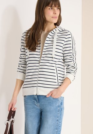 Frau trägt eine weiße Kapuzenjacke mit durchgehendem Reißverschluss und schwarzen Streifen, hellblaue Jeans, hält einen braunen Handtaschengurt und hat eine Hand in der Tasche.