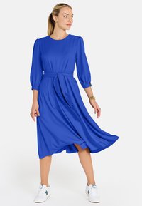 Žena v plynúcej midi šatách kráľovskej modrej farby s tromi štvrtinovými rukávmi a bielymi teniskami, pózujúca na jednoduchom svetlom pozadí.