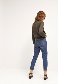 Sweter z dzianiny w kolorze oliwkowym z fakturą, dżinsy w niebieskim kolorze z wysokim stanem, czarne sandały na słupku, widoczne z tyłu na jednolitym tle.