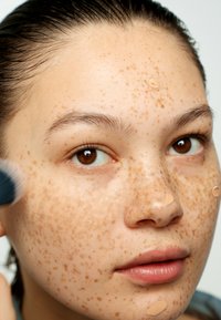 Gros plan sur un visage avec un léger hâle et de nombreuses taches de rousseur. Des points de fond de teint sont appliqués sur la peau, avec un pinceau à maquillage dans le cadre.