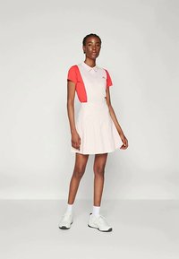 Polo-Kleid mit rosem Oberteil und weißem Rock, das ein plissiertes Design aufweist. Hergestellt aus einem glatten Stoff und getragen mit weißen Sneakern.