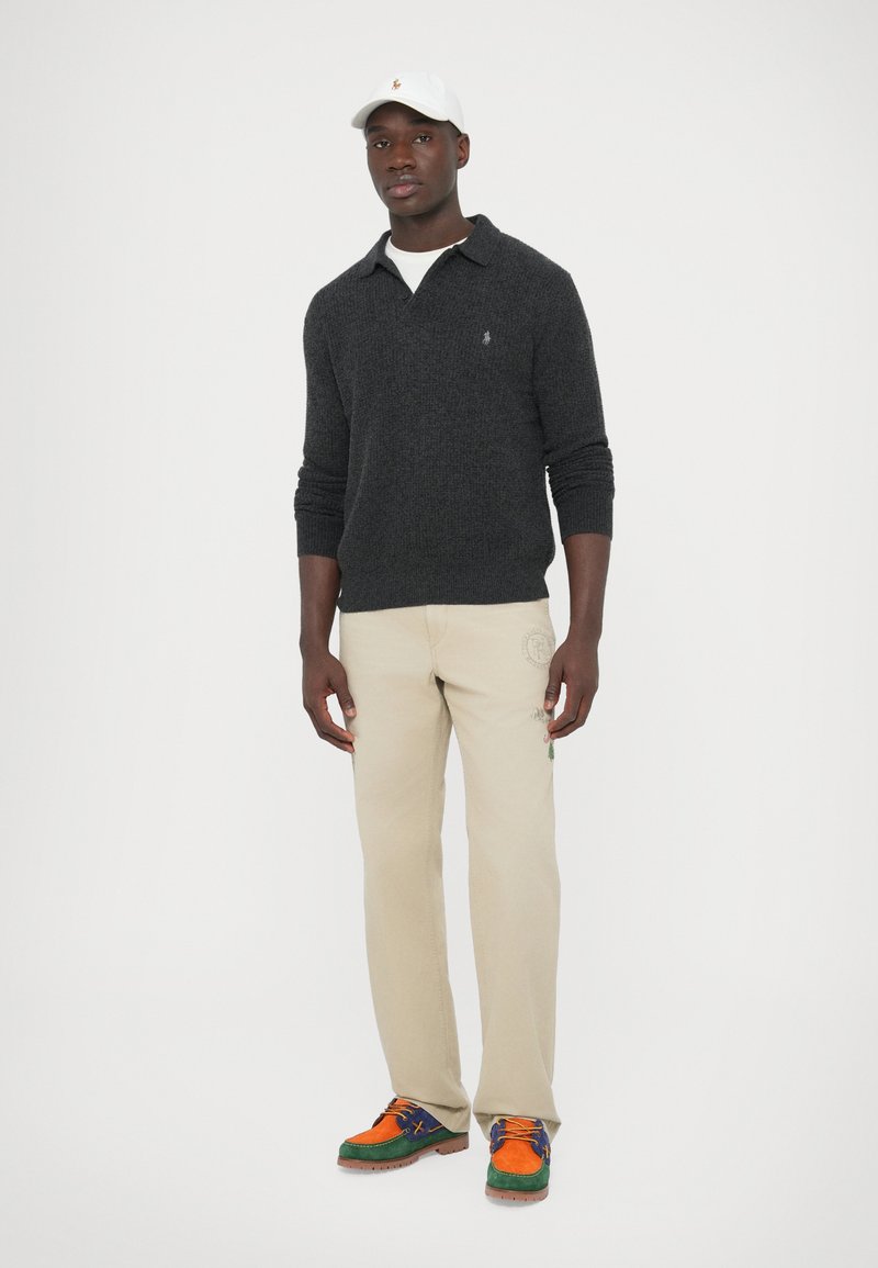 Maglione lavorato a maglia grigio scuro, pantaloni beige con dettagli del logo e sneakers multicolori con accenti arancioni, blu e verdi. Cappellino bianco.