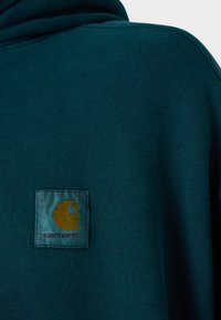 Dunkelblaue Sweatshirt mit weicher Textur und Kapuze. Verfügt über ein quadratisches Carhartt-Logo-Patch in Gold und Rot auf der linken Brustseite.