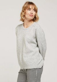 Pull en maille gris clair avec un col rond et des poignets côtelés, offrant une coupe décontractée. Porté avec un pantalon à carreaux noir et blanc.